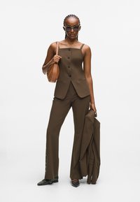 Woman with braided hair wearing brown buttoned sleeveless top and matching pants, holding a brown jacket and leather handbag.