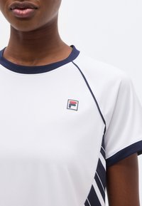 Κοντινό πλάνο ενός ατόμου που φοράει αθλητική μπλούζα σε λευκό και ναυτικό χρώμα με μικρό λογότυπο FILA στο στήθος και πλευρικά πάνελ με ρίγες σε ναυτικό χρώμα.