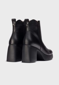 Botas de tobillo de cuero negro con punta redondeada y cuello con forma de ondas. Tienen un tacón grueso y mate, y cierre con cremallera en la parte posterior.