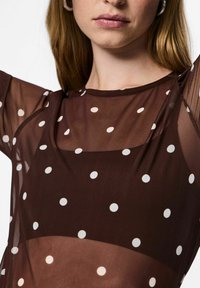 Primo piano di una donna che indossa una maglietta trasparente marrone a pois con pois bianchi e orecchini a cerchio, con le braccia sollevate fuori dal campo.