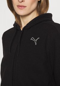 Felpa nera con zip, caratterizzata da una tasca a marsupio, cappuccio con coulisse e logo ricamato bianco sul lato sinistro del petto. Texture morbida.