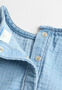 Helle blaue Denim-Stoff mit strukturierter Oberfläche, ausgestattet mit einem geknöpften Kragen und Metall-Druckknöpfen. Markenlabel innen sichtbar.