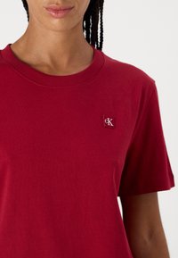 Μπορντό βαμβακερό t-shirt με κανονικό γιακά, κοντά μανίκια και μια μικρή ετικέτα CK στο στήθος. Λείο υλικό και κλασική εφαρμογή.