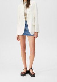 Kremowa marynarka z dwoma guzikami, połączona z jasnym dopasowanym topem, dżinsową mini spódnicą oraz czarnymi platformami na paski wokół kostki.