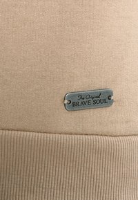 Tessuto beige a maglia con orlo a coste e targhetta metallica incisa con "The Original BRAVE SOUL." La texture è morbida e liscia.