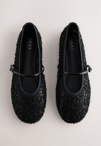 Chaussures Mary Jane noires à sequins avec fines brides réglables, semelles plates et bouts arrondis, présentées côte à côte sur un fond clair.