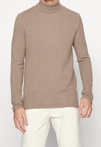 Beige högt halsad tröja bärs av person med krämfärgade byxor, som visar överkropp och armar mot en neutral bakgrund.