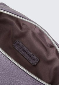 Interno di una borsa in pelle viola con foderatura liscia marrone scuro e un'etichetta con logo cucita. Dotata di una cerniera metallica.