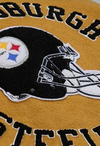 Schwarz-weiß gestickter Plüsch-Fußballhelm auf goldenem Stoffhintergrund mit dem Pittsburgh Steelers Logo und drei farbigen Sternen.