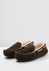 Pantofole in suede marrone con morbida fodera in pelliccia beige, punta rotonda e cucitura decorativa lungo il bordo. Suola in gomma per una migliore aderenza.