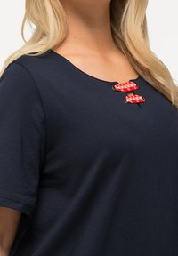 Tmavě modré tričko s kulatým výstřihem, zdobené třemi červenobílými kostkovanými akcenty na předním rozparku. Jemná, hladká textura.