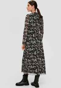 Langes, durchsichtiges Kleid in Schwarz mit grauem Leopardenmuster. Hat lange Ärmel und eine fließende Silhouette. Wird mit schwarzen Knöchelstiefeln getragen.