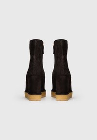 Un par de botines de ante negro con patrón texturizado y tacones en cuña beige, vistos desde atrás sobre un fondo blanco.