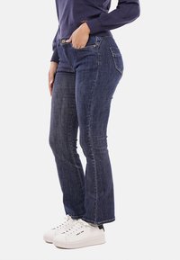 Jeans a campana in denim scuro con cinque tasche, patta con zip e accessori in ottone, abbinati a sneakers bianche e un top a maniche lunghe blu navy.
