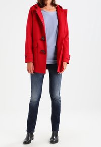 Rode wollen jas met klittenbandsluitingen, twee voorzakken en een capuchon. Gecombineerd met een lichtblauw shirt en donkerblauwe spijkerbroek, aangevuld met zwarte laarzen.