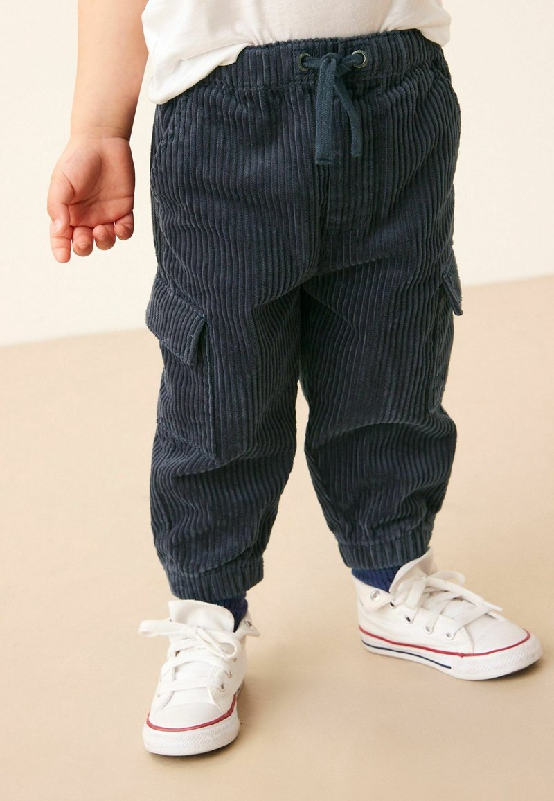 Tout-petit portant un pantalon en velours côtelé bleu foncé avec des poches latérales, des baskets blanches et un haut blanc, debout sur un sol beige.