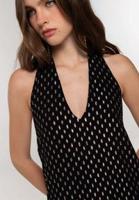 Giovane donna con lunghi capelli castani che indossa un vestito nero a halter con motivi rettangolari dorati e orecchini d'oro pendenti.