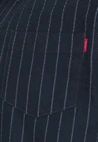 Navy fabric shirt with white vertical stripes, featuring a single chest pocket and a small red logo tag on the side. Textured cotton material.