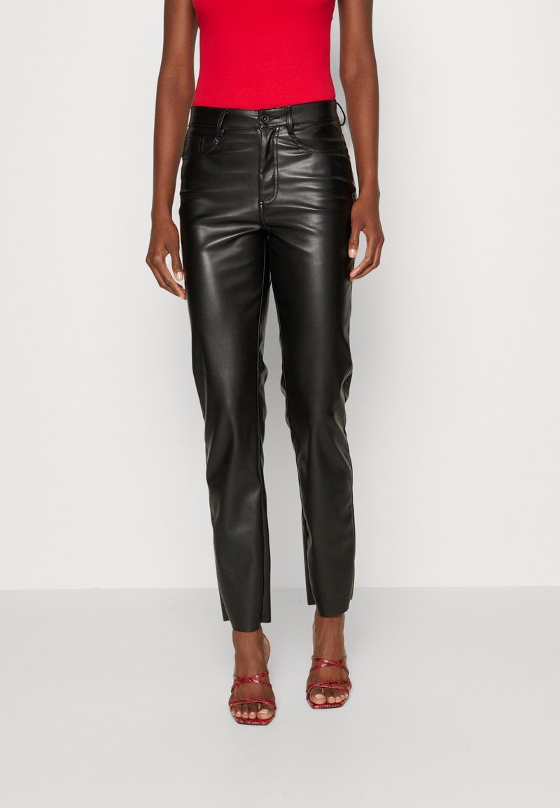 Personne portant un pantalon en cuir noir, un haut rouge sans manches et des sandales rouges à talons hauts à brides, debout devant un fond uni.