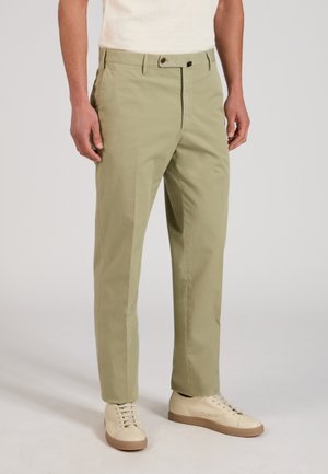 Mężczyzna noszący proste spodnie w kolorze khaki i kremowo-białe trampki, stojący z rozluźnionymi rękami swobodnie opuszczonymi wzdłuż boków na jednolitym tle.