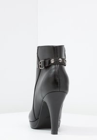 Czarne skórzane botki za kostkę z ostrym noskiem, wysokim obcasem oraz ozdobną klamrą z piramidowymi ćwiekami wzdłuż pasków za kostkę. Gładka tekstura.
