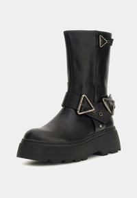 Stivale alla caviglia in pelle nera con una suola piatta spessa, caratterizzato da fibbie decorative a forma triangolare e una texture liscia.