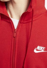 Person, der einen roten Nike-Reißverschluss-Hoodie mit einem weißen Logo auf der Brust trägt und ein teilweise sichtbares weißes T-Shirt darunter hat.