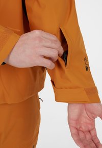 Narančasta vodootporna jakna s patentnim zatvaračem na rukavu i čičak manžetama. Glatka tekstura, funkcionalan dizajn, prikladna za vanjsku upotrebu.