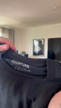 Juodas medvilnės marškinėliai su apvaliu kaklu, lygia tekstūra ir etiketėmis, žyminčiomis dydžius nuo S iki XXL. Matoma prekės ženklas "YOURTURN".