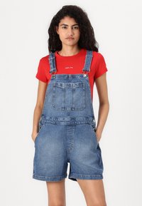 Macacão de ganga com um tom azul claro, apresentando alças ajustáveis, dois bolsos frontais e um corte largo, combinado com uma camisa de mangas curtas vermelha.