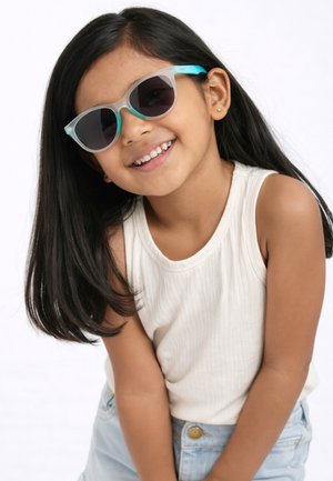 Joven chica de cabello largo y oscuro, que lleva gafas de sol en tonos verde azulado y gris, una camiseta blanca sin mangas y vaqueros azul claro, sonriendo sobre un fondo blanco.