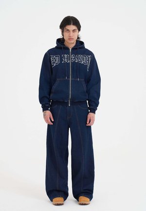 Jeans-Zip-Hoodie mit "ED HARDY" Schriftzug in Weiß, kombiniert mit weit geschnittenen Jeanshosen. Beide Teile zeichnen sich durch kontrastierende Nähte und einen lässigen Schnitt aus.