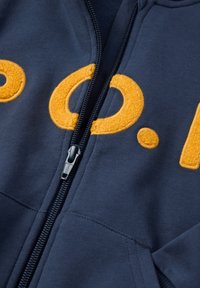 Marineblauwe rits-hoodie met grote, gestructureerde oranje letteren. Glad materiaal met ribbeldetails bij de rits en zakken.