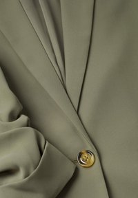 Veste blazer vert olive avec un seul bouton en écaille de tortue et un détail de tissu froncé sur la manche.