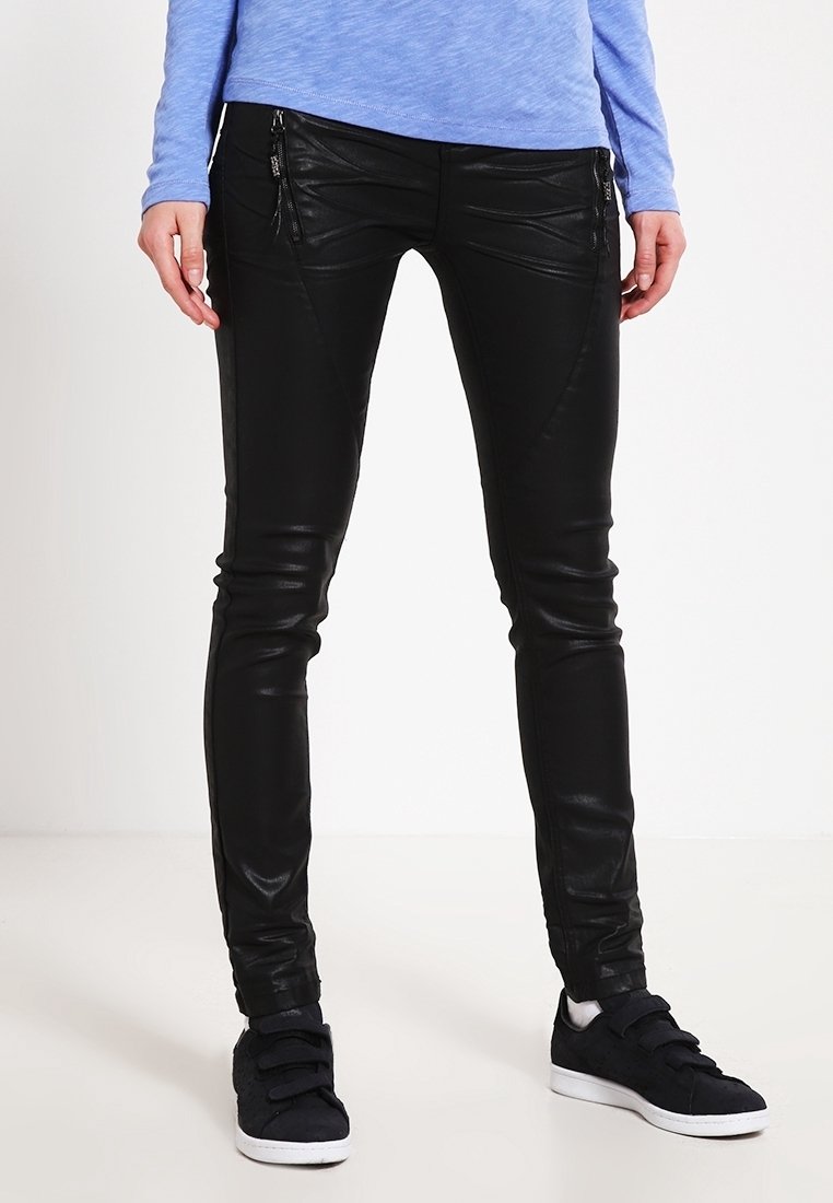 Personne portant un pantalon en cuir noir slim avec des détails de fermeture éclair sur le devant et des baskets noires à semelles blanches, debout devant un fond uni.