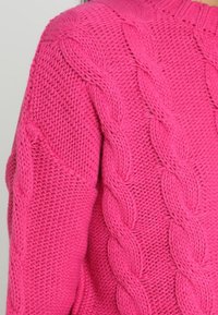 Manche de pull épais en tricot rose vif avec un grand motif torsadé tressé et un détail de point texturé.