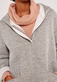 Szara bluza z zamkiem błyskawicznym i białą podszewką oraz różowy sweter z dzianiny z okrągłym dekoltem pod spodem. Posiada ściągacze przy mankietach i kieszenie.