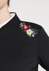 Černá mikina s vysokým límcem, zdobená vyšitými květinovými detaily. Design zahrnuje červené a bílé květiny s zelenými listy.
