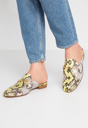 Person iført blå jeans og slip-on loafers med gult og gråt slangeskindsmønster og små træhæle.