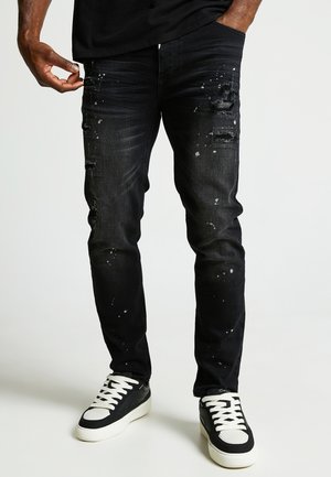 Jeans in denim nero con una vestibilità slim, dettagli strappati, macchie di vernice e bordi sfrangiati, abbinati a sneakers nere.