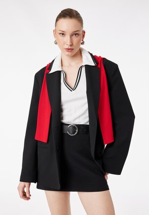 Młoda kobieta z zaczesanymi do tyłu włosami, ubrana w czarną marynarkę na białą koszulę z czarną lamówką, czarną spódnicę i czerwoną tkaninę narzuconą na ramiona.