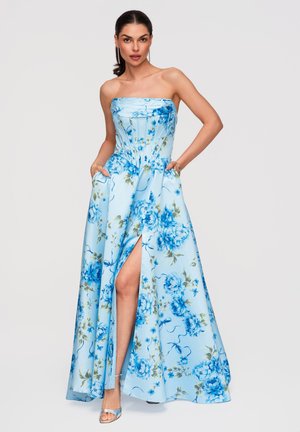 Kobieta w jasnoniebieskiej sukni bez ramiączek z niebieskimi kwiatowymi wzorami i rozcięciem do uda, ręce w kieszeniach oraz srebrne sandały na obcasie z odkrytymi palcami.