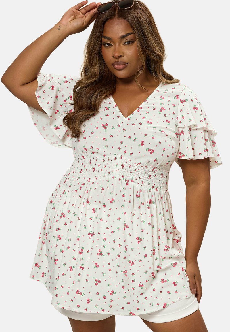 Kobieta o długich, falowanych włosach, ubrana w białą, kwiecistą sukienkę z falbanami na rękawach, trzymająca okulary przeciwsłoneczne na głowie na jednolitym tle.