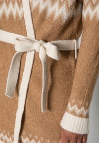 Virkad cardigan i ljusbrun färg med ett vitt zickzackmönster i nederkant och på ärmarna. Har ett krämfärgat knytbälte, ribbade ärmslut och en mjuk textur.