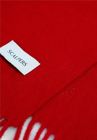 Bufanda de lana roja con una etiqueta de tela blanca etiquetada "SCALPERS" y un diseño de calavera en relieve. Con flecos distintivos en uno de los bordes.
