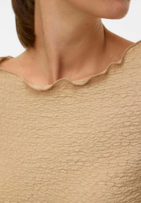 Primo piano del collo e della parte superiore del torace di una persona che indossa un top beige con una texture e un girocollo ondulato.