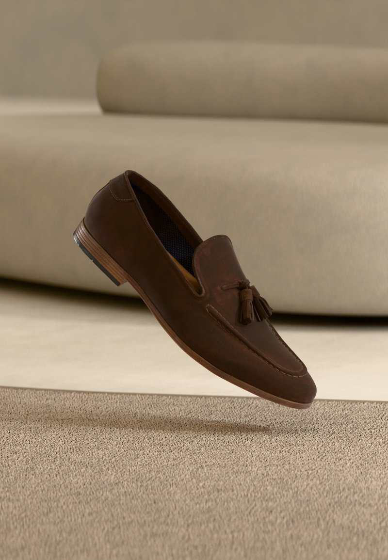 Mocassin en cuir marron avec des pampilles, flottant au-dessus d'un tapis beige et d'un canapé dans un intérieur doucement éclairé.