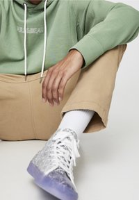 Felpa verde con cordini bianchi, pantaloni beige e sneakers con un motivo e suola traslucida. Calzini bianchi che sbucano sopra le scarpe.
