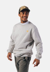 Jasnoszary sweatshirt z ribbowe rękawami i dołem, z małym logo żółtego słonia. W połączeniu z szarymi spodniami i beżową czapką.