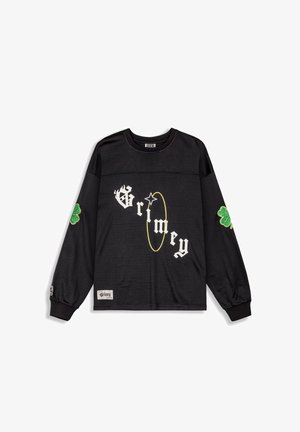Camisa de manga larga negra con textura de malla, con el texto blanco "Grimey" y un acento en círculo amarillo. Parche de trébol verde en las mangas.